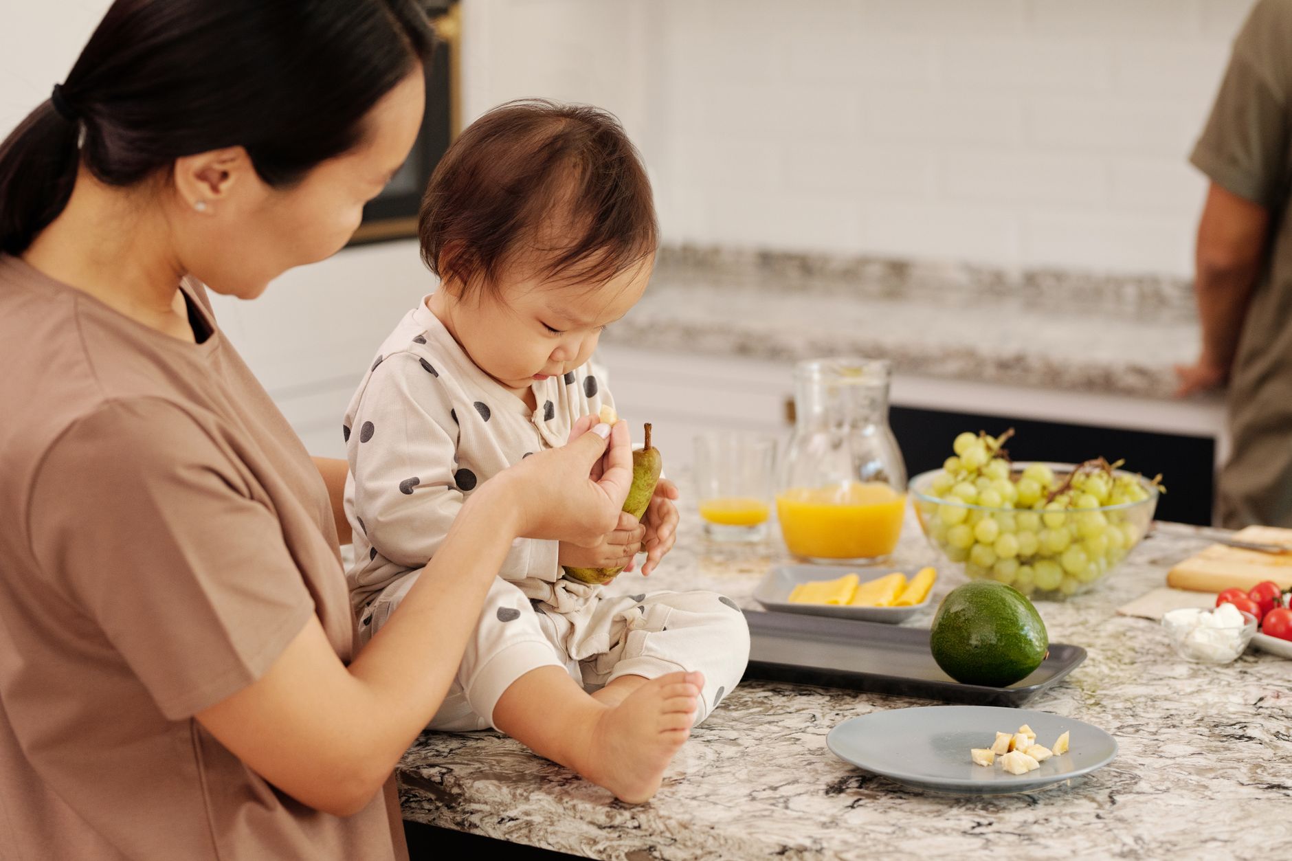 An adult feeding fruit to a baby at a kitchen counter. Stock photograph.