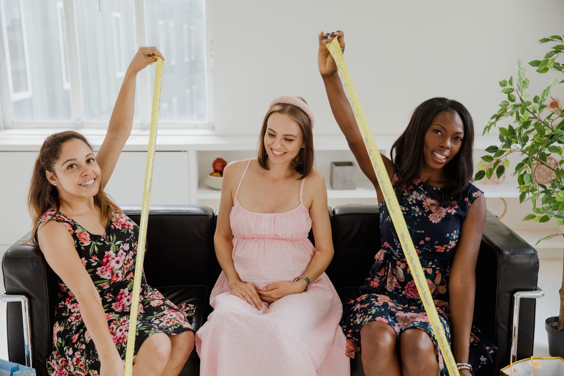 Three people sitting together on a sofa, one of them pregnant, laughing over a tape measure. Stock photograph.